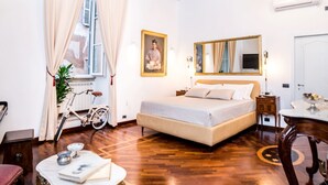 Ein geräumiges Zimmer mit einem großen Bett, einem Schreibtisch und einem Stuhl. Ein Fenster mit Vorhängen, eine Kristallleuchter und ein Gemälde an der Wand sind ebenfalls vorhanden.