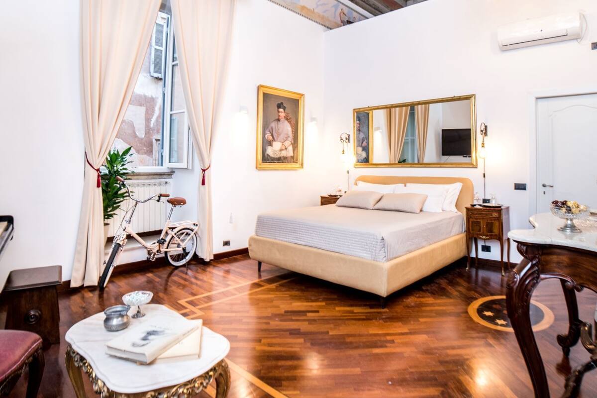 Una habitación amplia con una cama grande, un escritorio y una silla. Cuenta con un ventanal con cortinas, un candelabro y un cuadro en la pared.