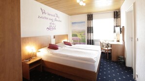 Ein Hotelzimmer mit Holzbalkendecke, einem großen Bett mit weißer Bettwäsche, zwei Nachttischen mit Lampen, einem Schreibtisch mit Stuhl und einem Fenster mit Blick auf Felder.