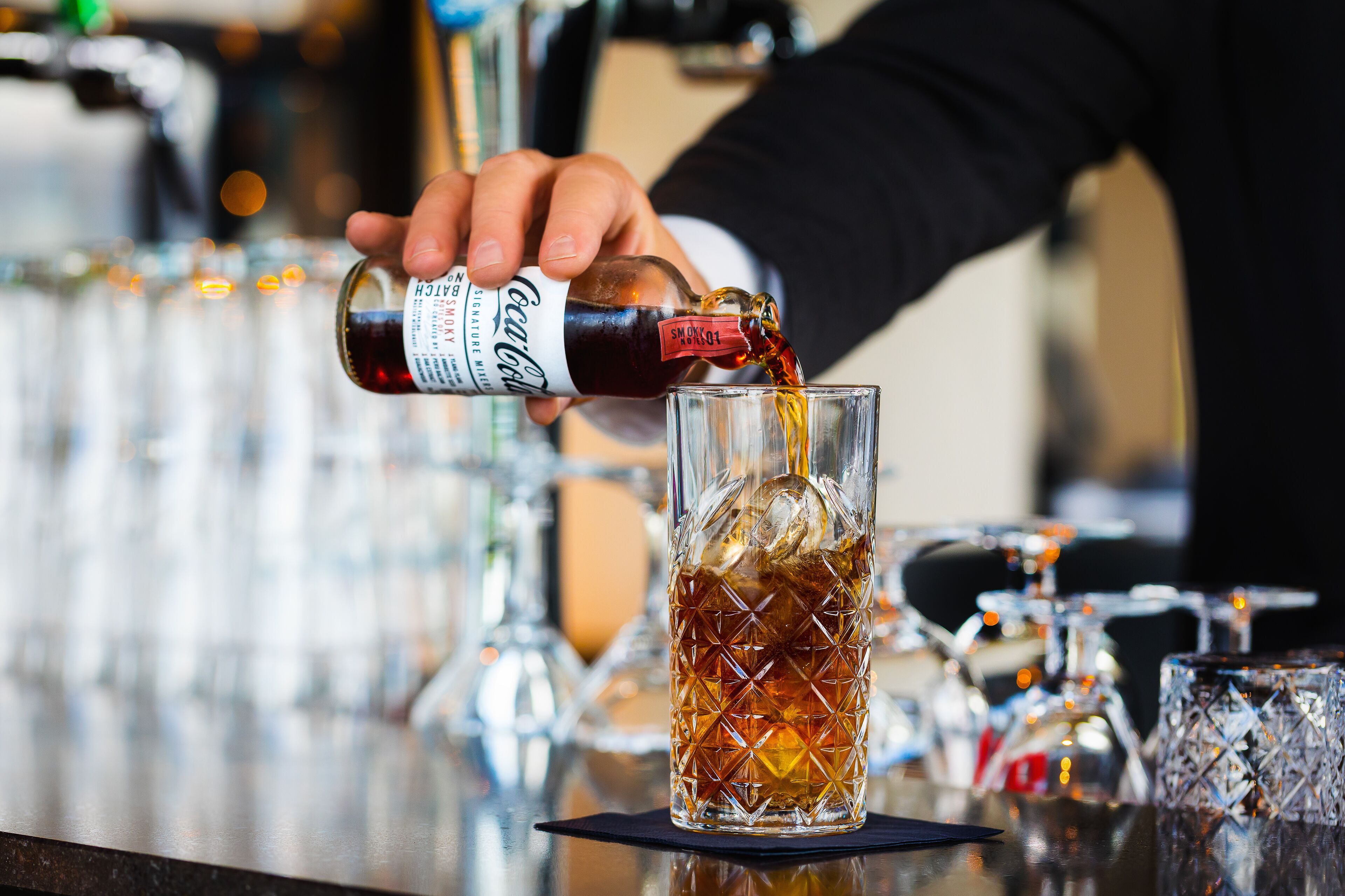 A hand pouring a dark liquid from a bottle into a glass with a textured pattern.