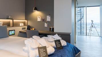 A modern hotel room with a large bed, a desk, and a view of the city through floor-to-ceiling windows.