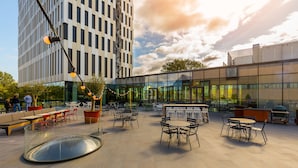 An outdoor seating area with tables and chairs, surrounded by modern buildings and a glass facade. - Postillion Hotel Amsterdam (Amsterdam)