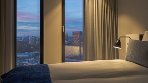A hotel room with a large bed, a view of the cityscape through the window, and a bedside lamp. - Postillion Hotel Amsterdam (Amsterdam)