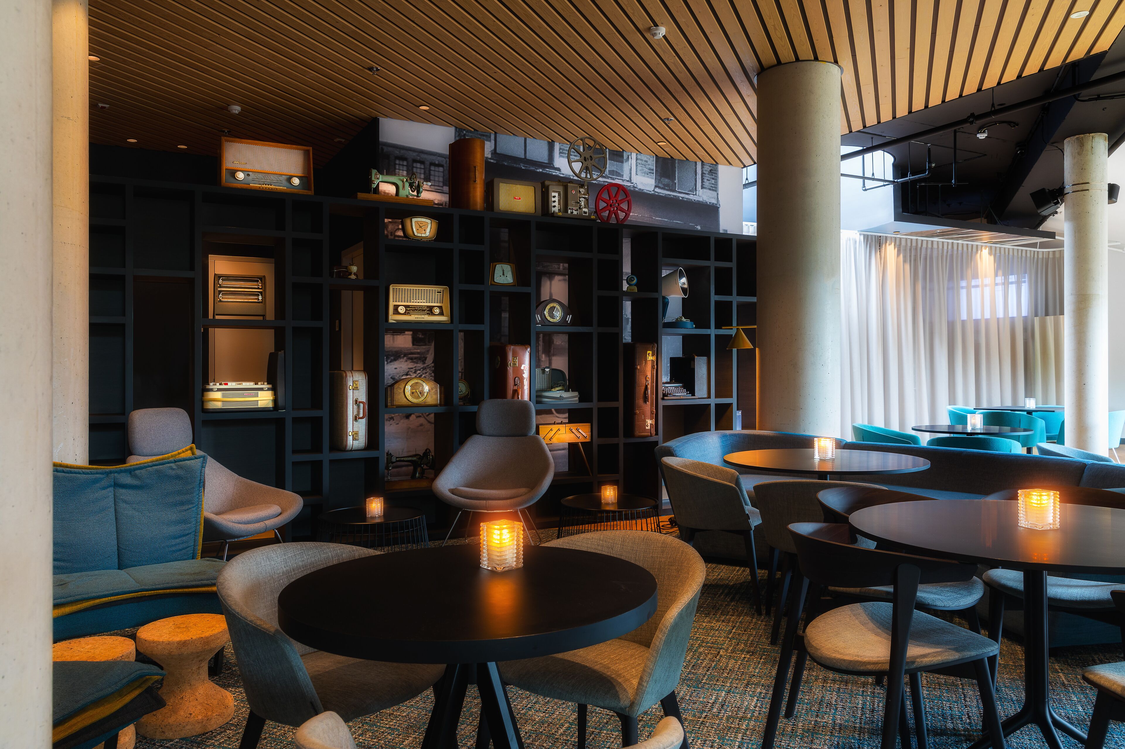 A modern lounge area with round tables, comfortable seating, and a dark wood-paneled ceiling.