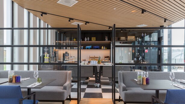 A modern restaurant with a checkered floor, grey seating, and a bar area visible through glass partitions. - Postillion Hotel Amsterdam (Amsterdam)