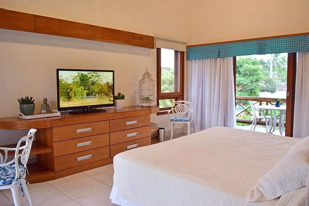Une chambre avec un lit, un téléviseur posé sur une commode en bois, une chaise et un balcon donnant sur un paysage verdoyant.