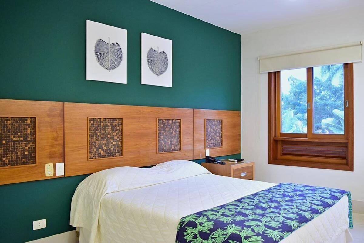 Une chambre à coucher comprenant un lit, une table de chevet, une fenêtre donnant sur des arbres et deux œuvres d’art accrochées au mur.