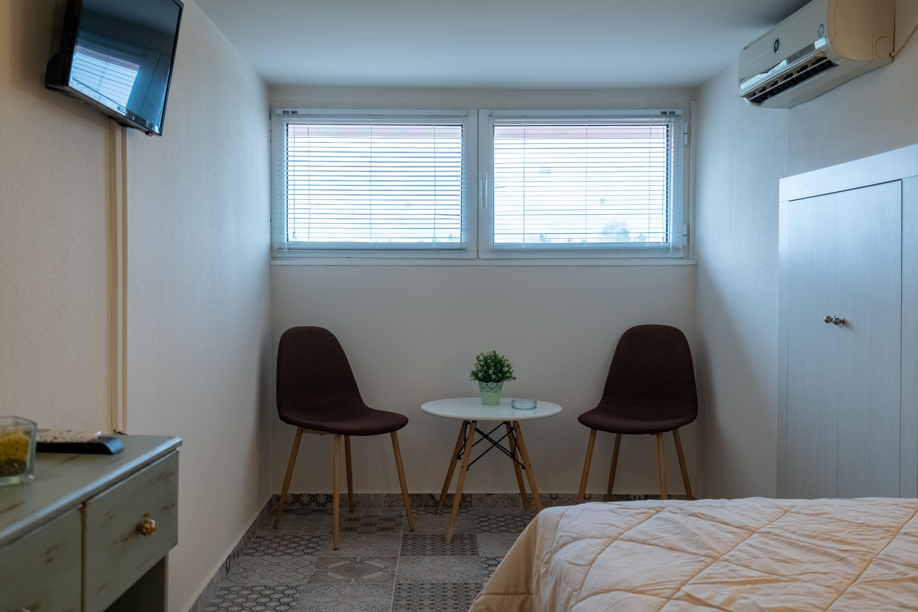 Um quarto pequeno com uma cama, uma escrivaninha, duas cadeiras, uma mesa de centro com uma planta e uma TV fixada na parede.