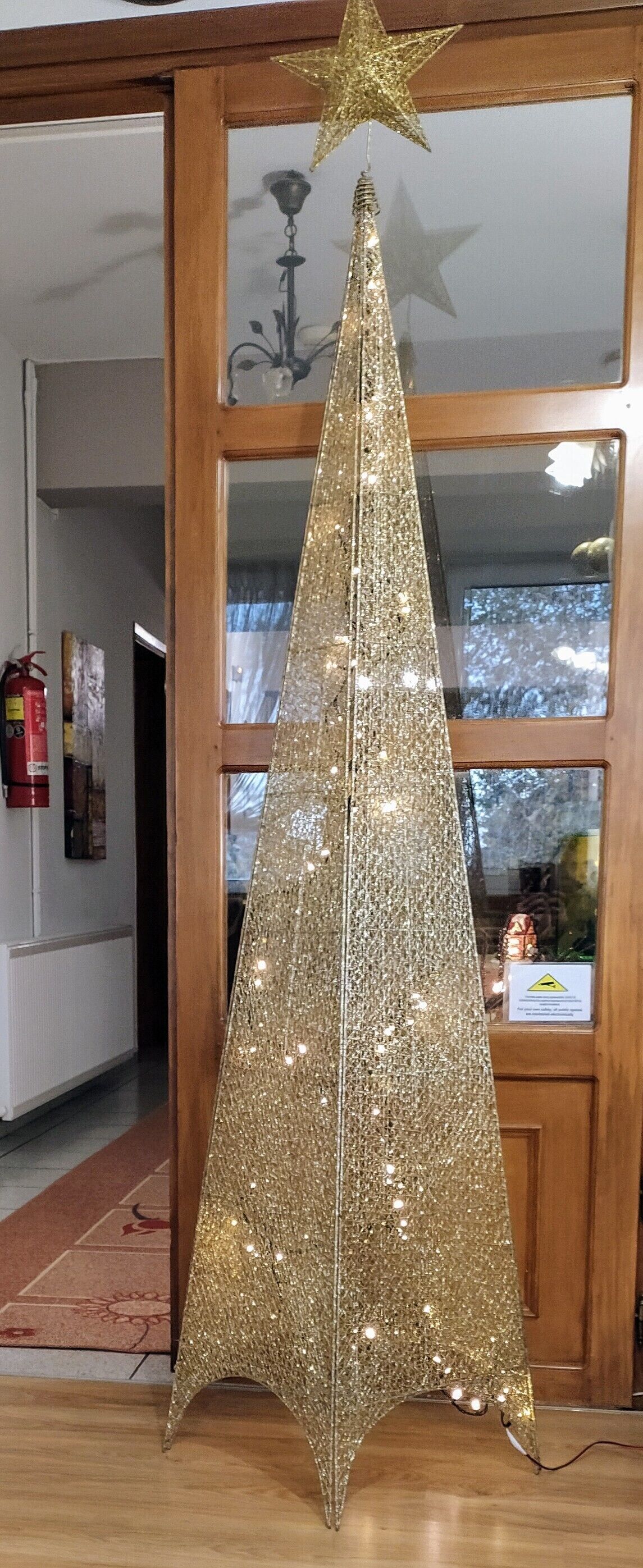A golden Christmas tree with lights in a room with wooden floors and a fire extinguisher on the wall.