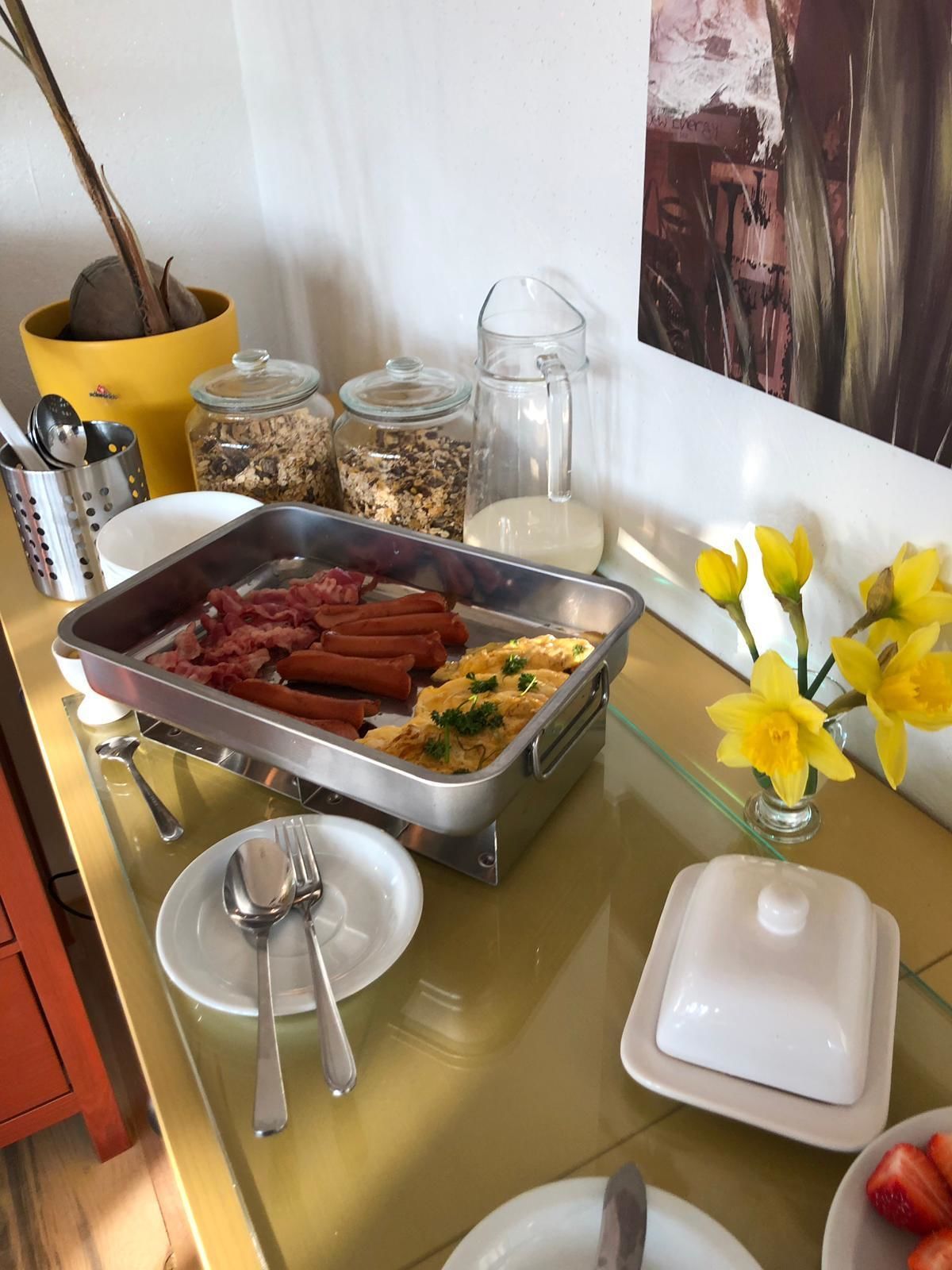Ein reichhaltiges Buffet mit Hot Dogs, Rührei und verschiedenen Beilagen. Außerdem befinden sich Blumen und eine Milchkaraffe auf dem Tisch.