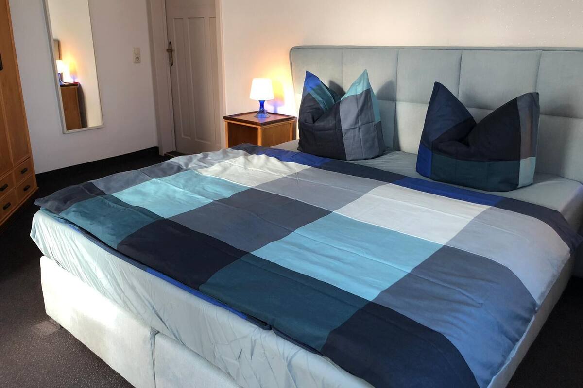Uma cama bem arrumada com um edredom azul e branco xadrez, uma cabeceira estofada cinza claro e duas luminárias de cabeceira.