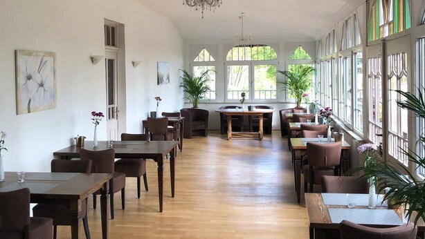 Un comedor amplio con mesas y sillas de madera, ventanas grandes y una araña.