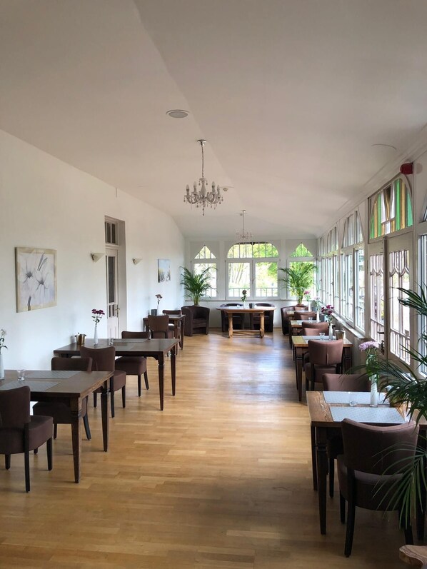 A spacious dining area with wooden tables and chairs, large windows, and a chandelier. - Hotel Zur Köppe (Bad Klosterlausnitz)