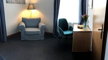 Et hotelværelse med en grå sofa, en turkis lænestol, et lille træbord med en vase blomster og en lampe på et sengebord.