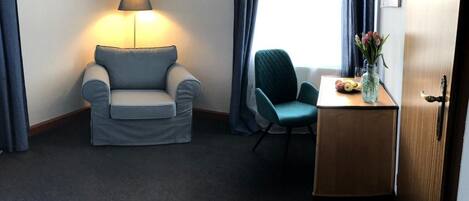 Habitación de hotel con un sofá gris, un sillón verde azulado, una pequeña mesa de madera con un jarrón de flores y una lámpara en una mesita.