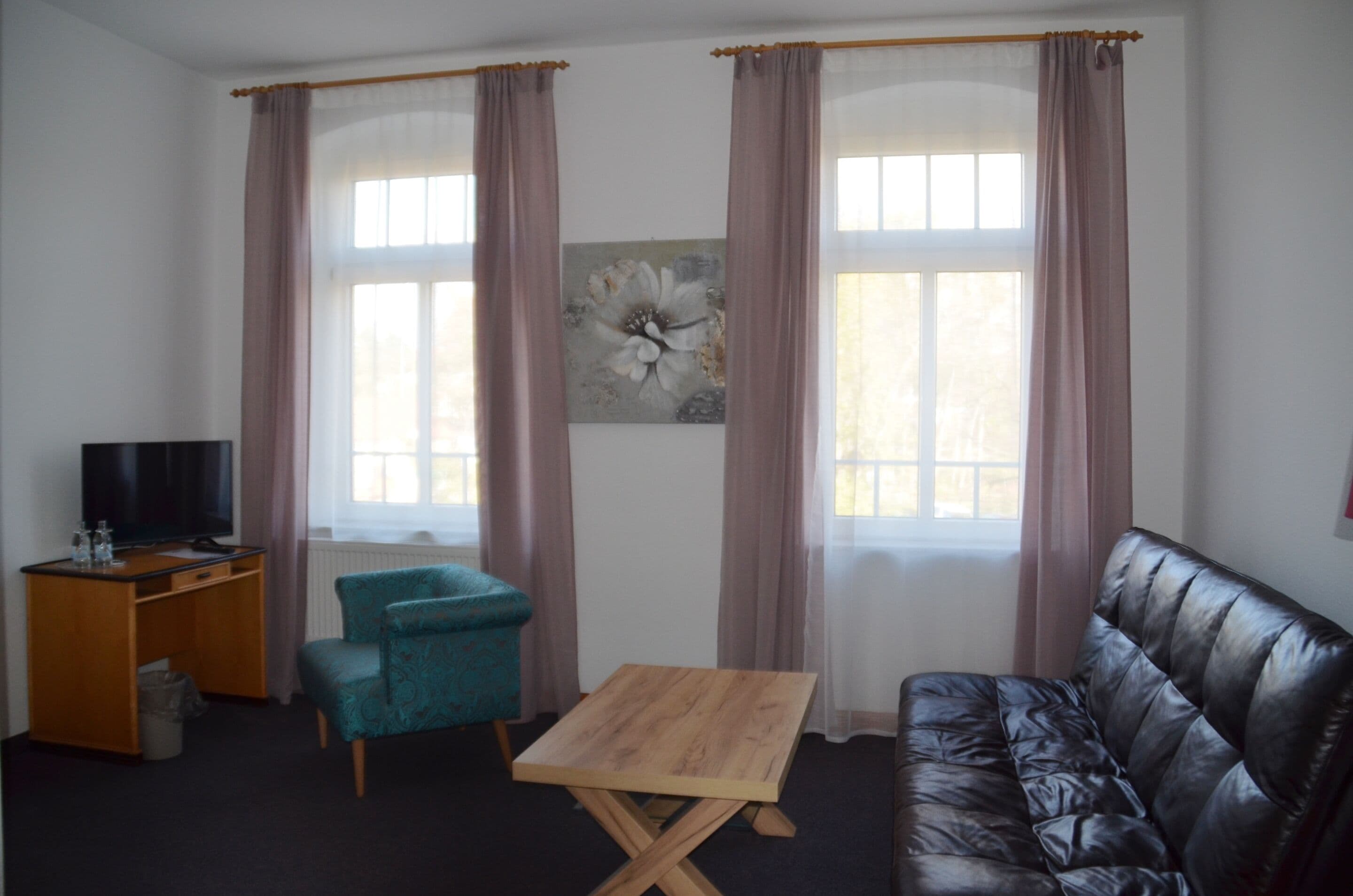 Un salon comprenant un canapé en cuir, une table basse en bois, un fauteuil turquoise, un bureau en bois avec une télévision et une grande fenêtre avec des rideaux.