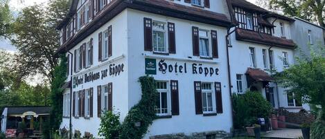 A traditional European hotel with a white facade and dark wooden shutters, featuring a sign for 'Hotel Kopp' and 'Biergarten.'