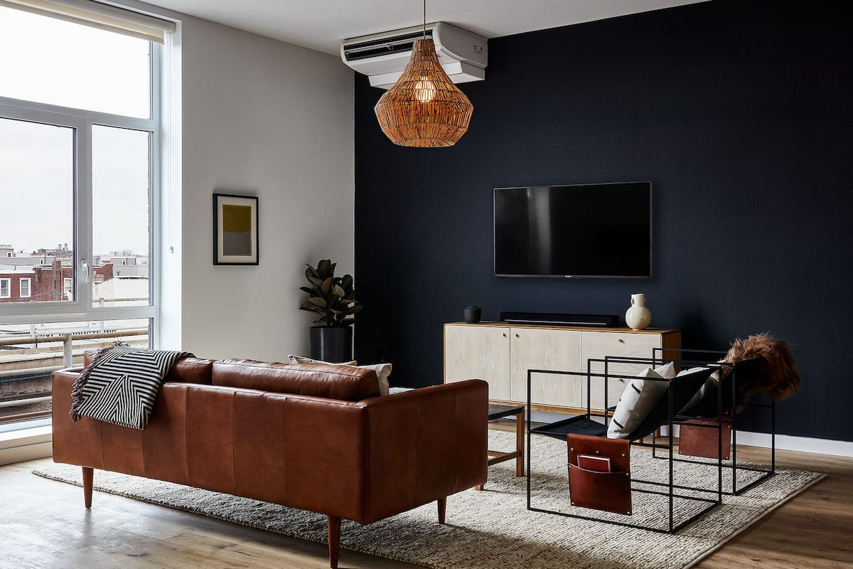 A modern living room with a large window, a brown leather sofa, a black TV, and a dark blue accent wall.