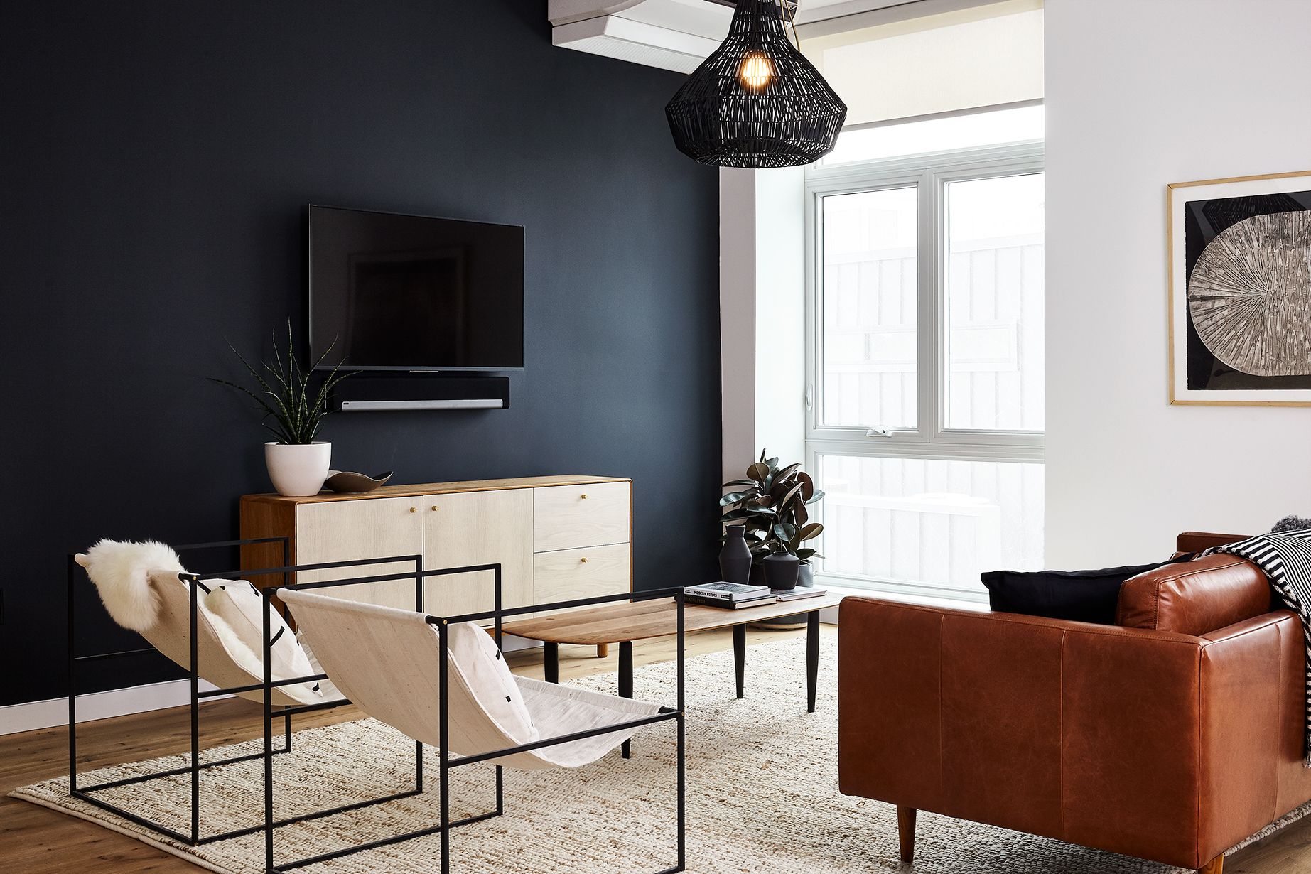 Ein modernes Wohnzimmer mit dunkler Wand, einem Flachbildfernseher, einer Holzkommode, einem Ledersofa und einer Pendelleuchte.