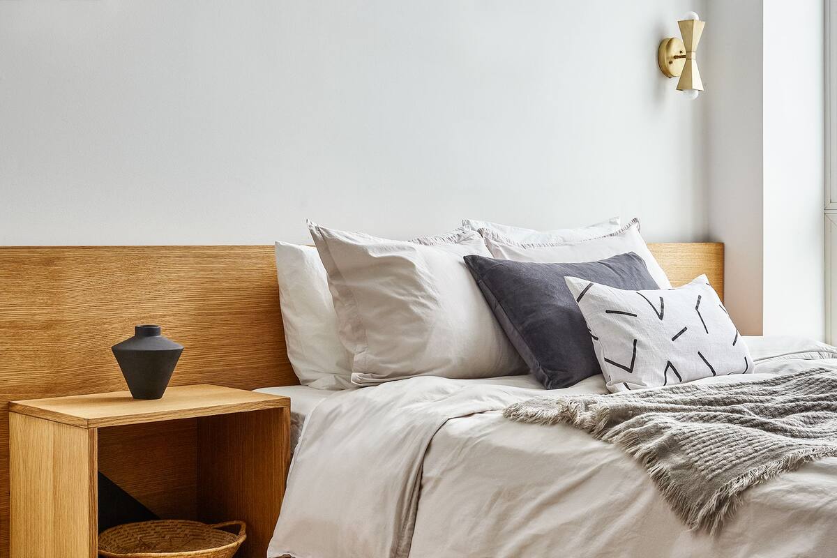 A neatly made bed with a wooden headboard, a bedside table with a vase, and a wall-mounted light fixture.