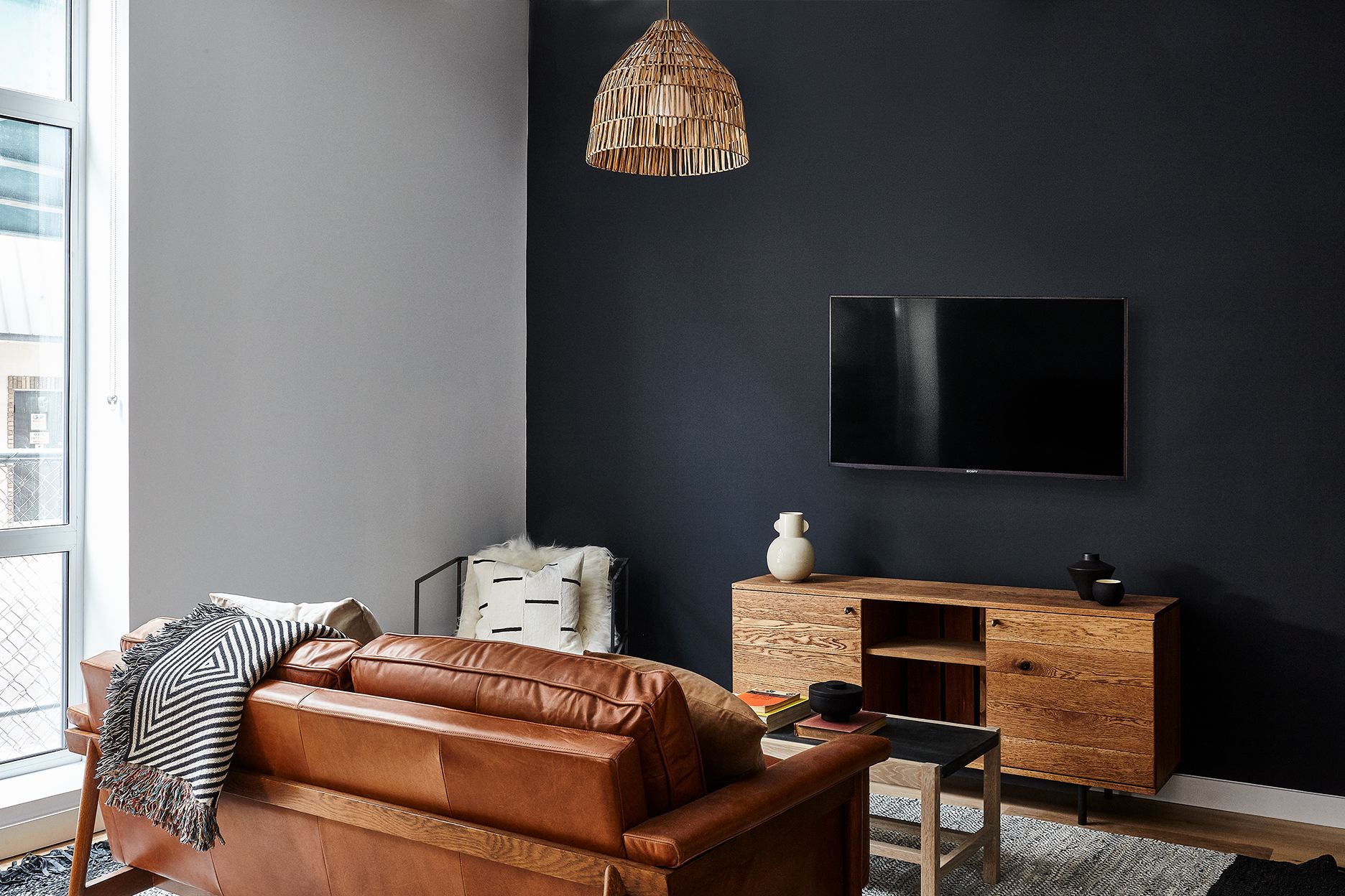 A modern living room with a dark wall, a leather sofa, a wooden cabinet, and a hanging light fixture.