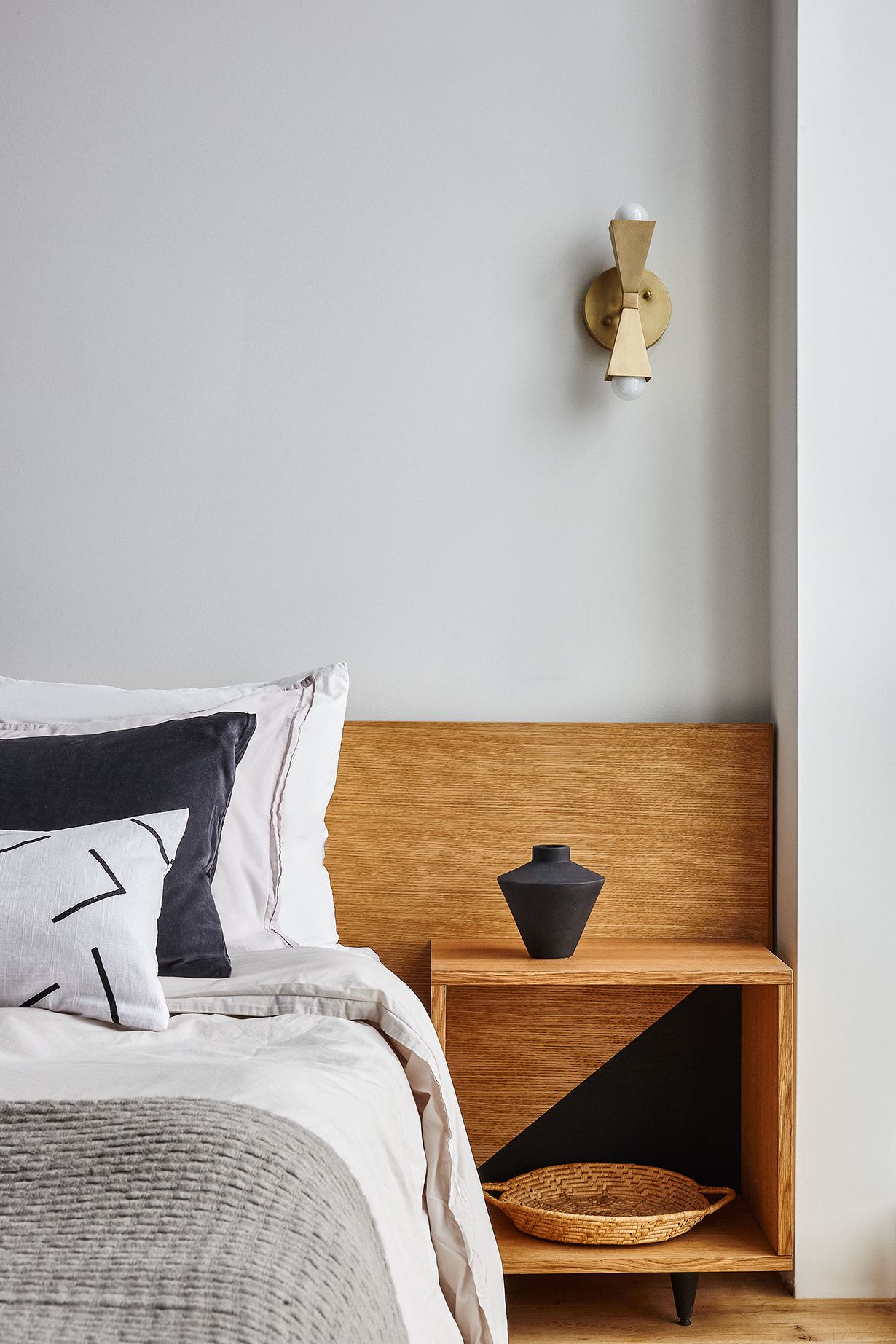 A neatly made bed with a grey and white duvet, a black pillow, and a grey knitted throw. A wooden nightstand with a black vase and a woven basket is beside the bed.