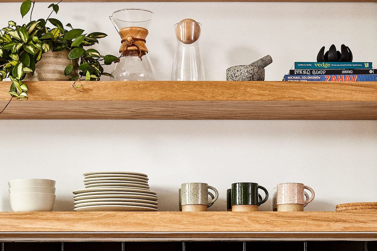 A kitchen with a white sink, black tiled backsplash, wooden cabinets, and a shelf with various kitchen items.