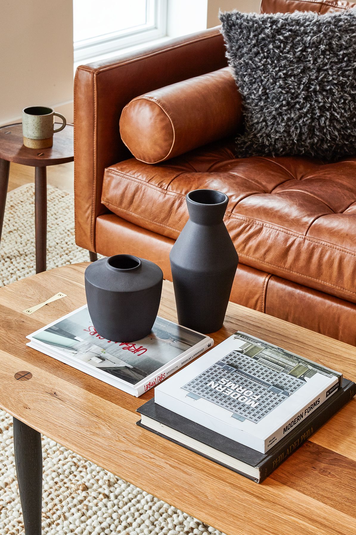 Um sofá de couro marrom com uma almofada cinza fofa, dois vasos pretos e dois livros sobre uma mesa de centro de madeira.