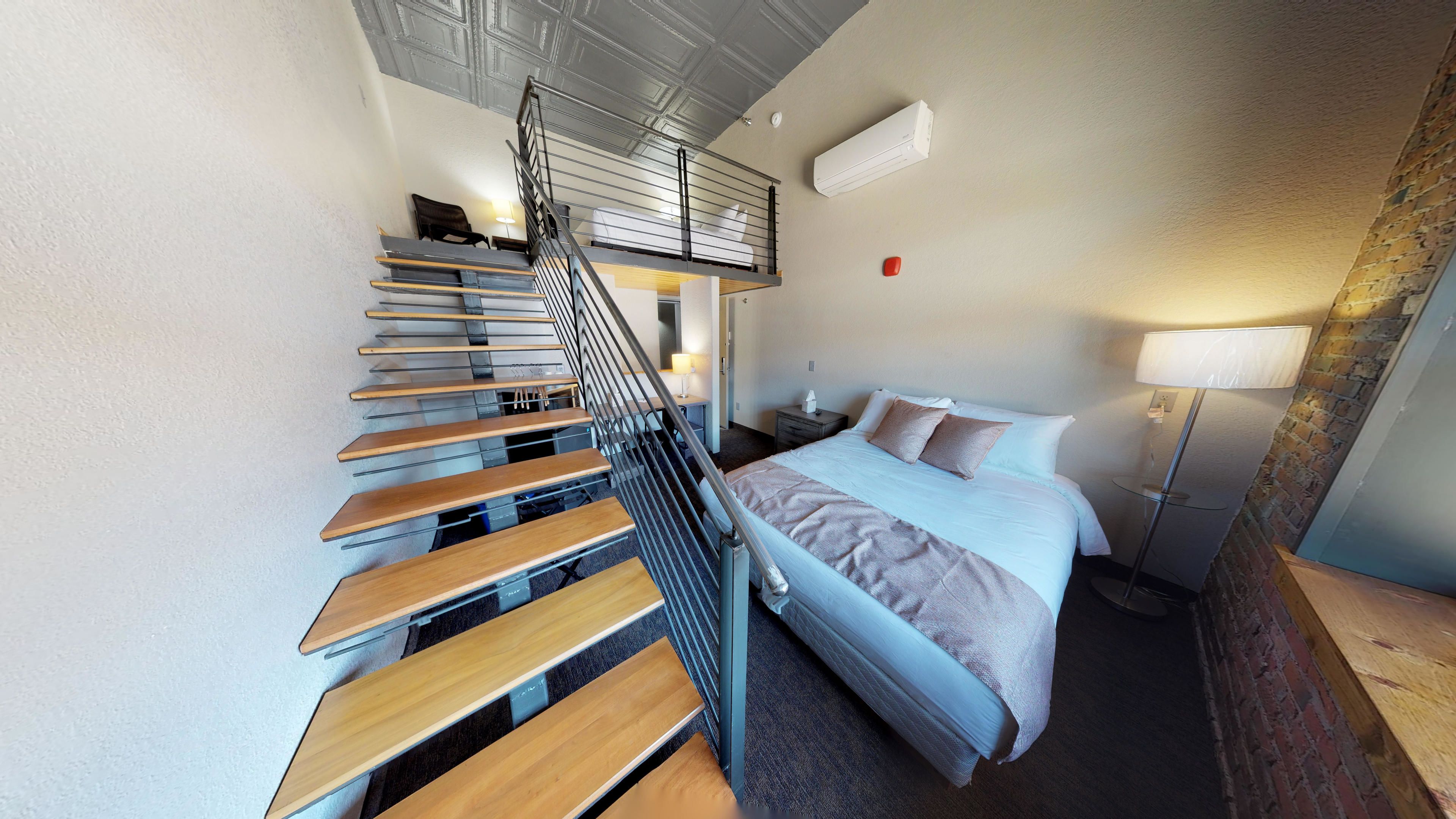 A modern hotel room with a wooden staircase, a bed with a white and brown duvet, a bedside table with a lamp, and a brick wall.