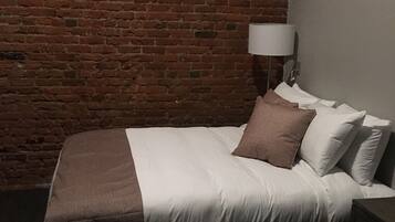 Un dormitorio con pared de ladrillo, una cama con ropa de cama blanca y marrón, una lámpara de noche y una mesita de noche de madera.