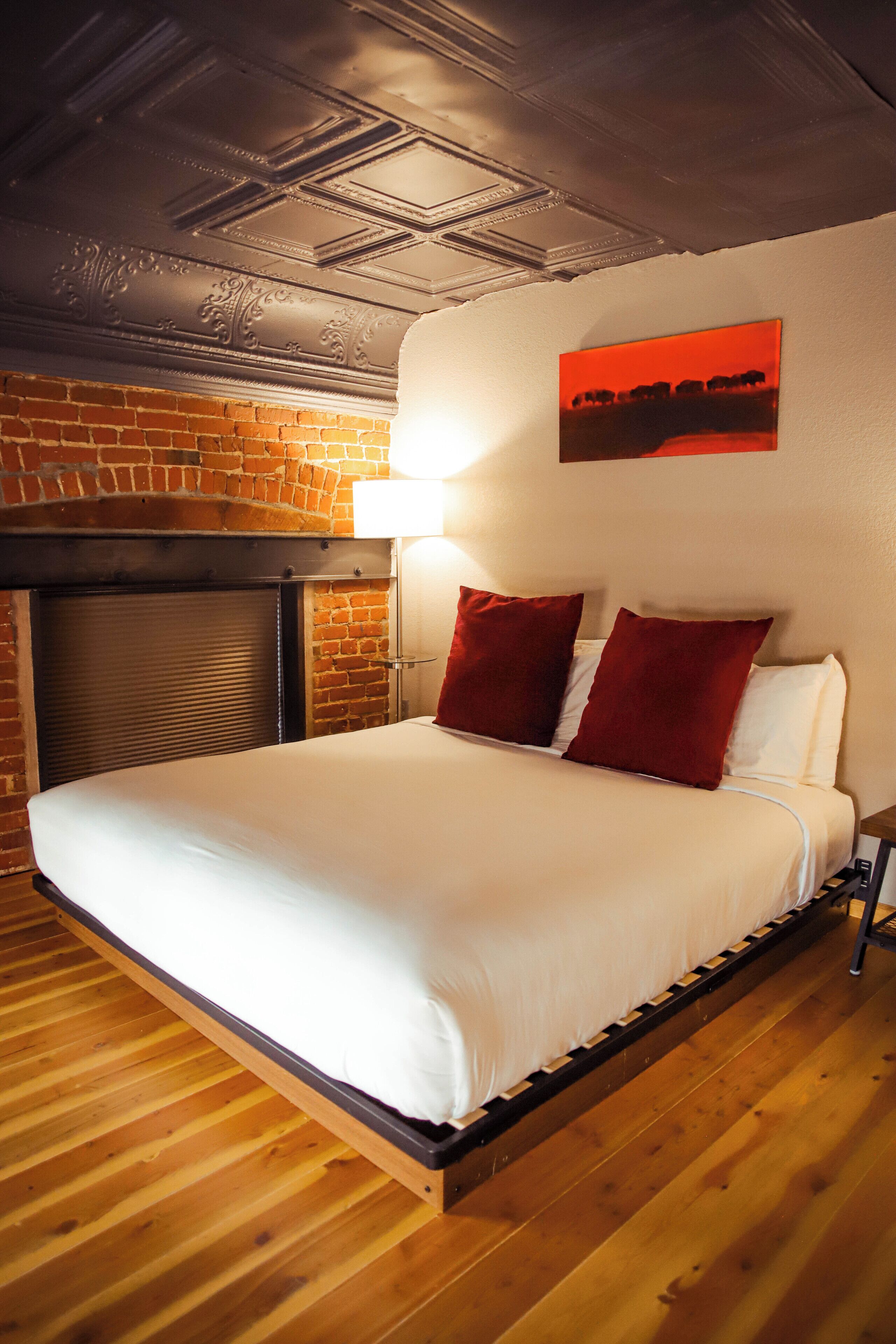 A hotel room with a brick fireplace, a bed with white bedding and red pillows, and a red painting on the wall.