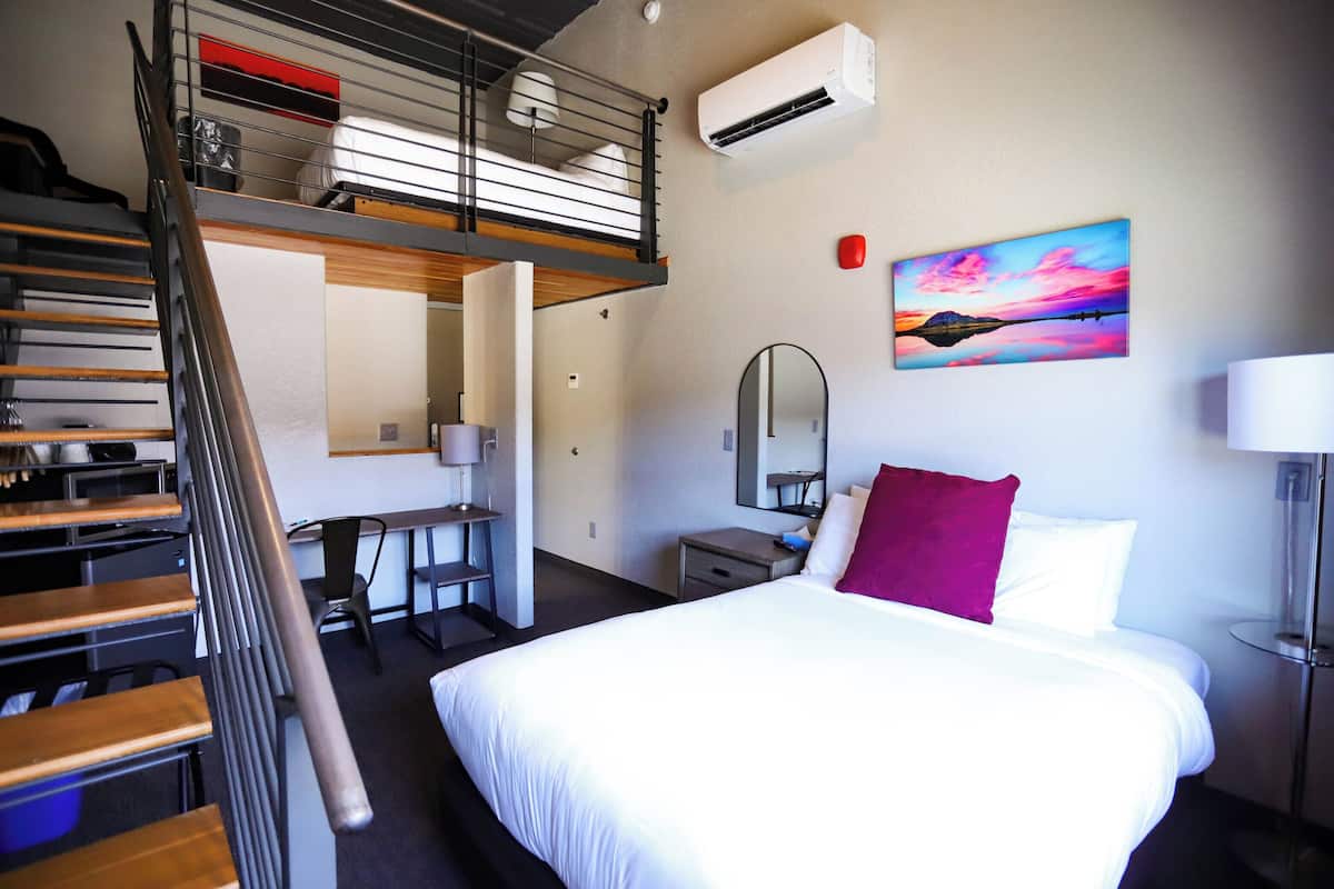A hotel room with a bed, a desk, a chair, a mirror, a staircase, and an air conditioning unit.