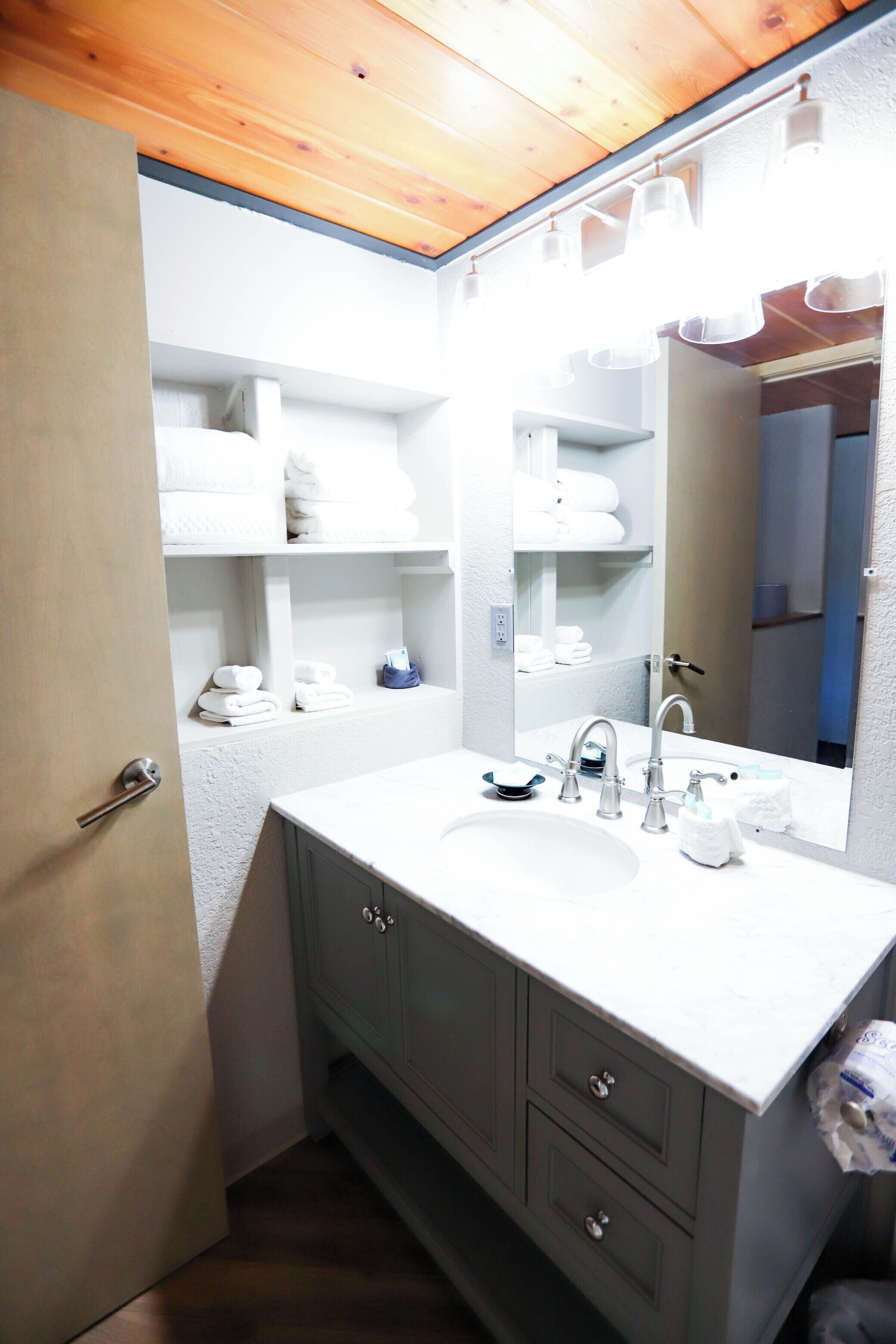 A bathroom with a white sink, two faucets, a mirror, and shelves with folded towels.