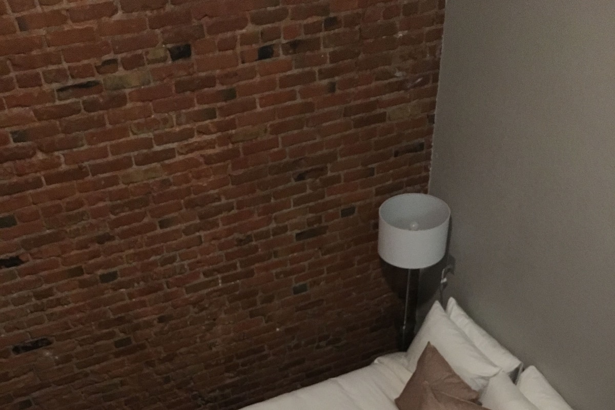 A single bed with white and brown bedding, a lamp, and a brick wall.