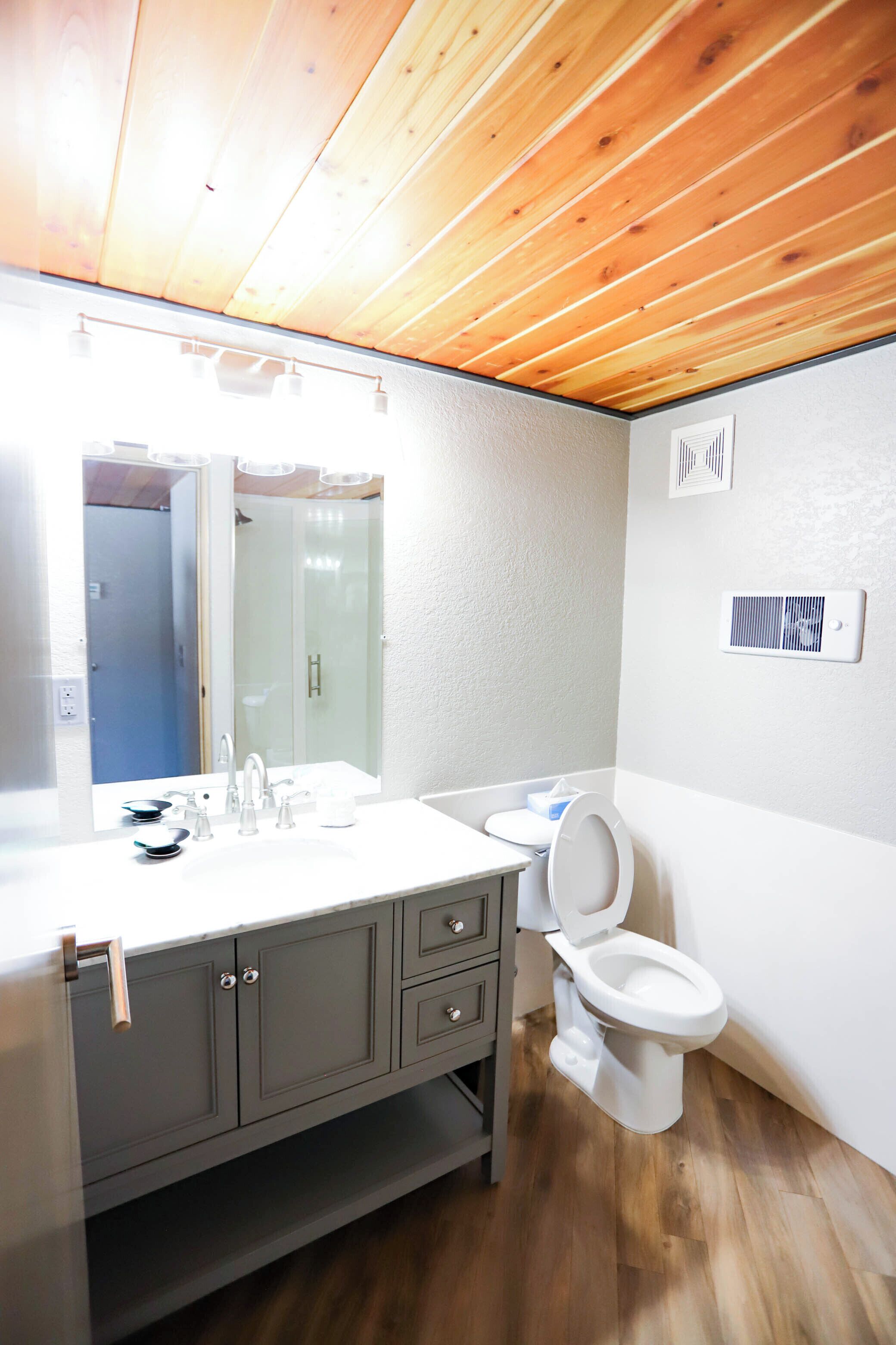 A bathroom with a wooden ceiling, a large mirror, a sink with a faucet, a toilet, and a shower.