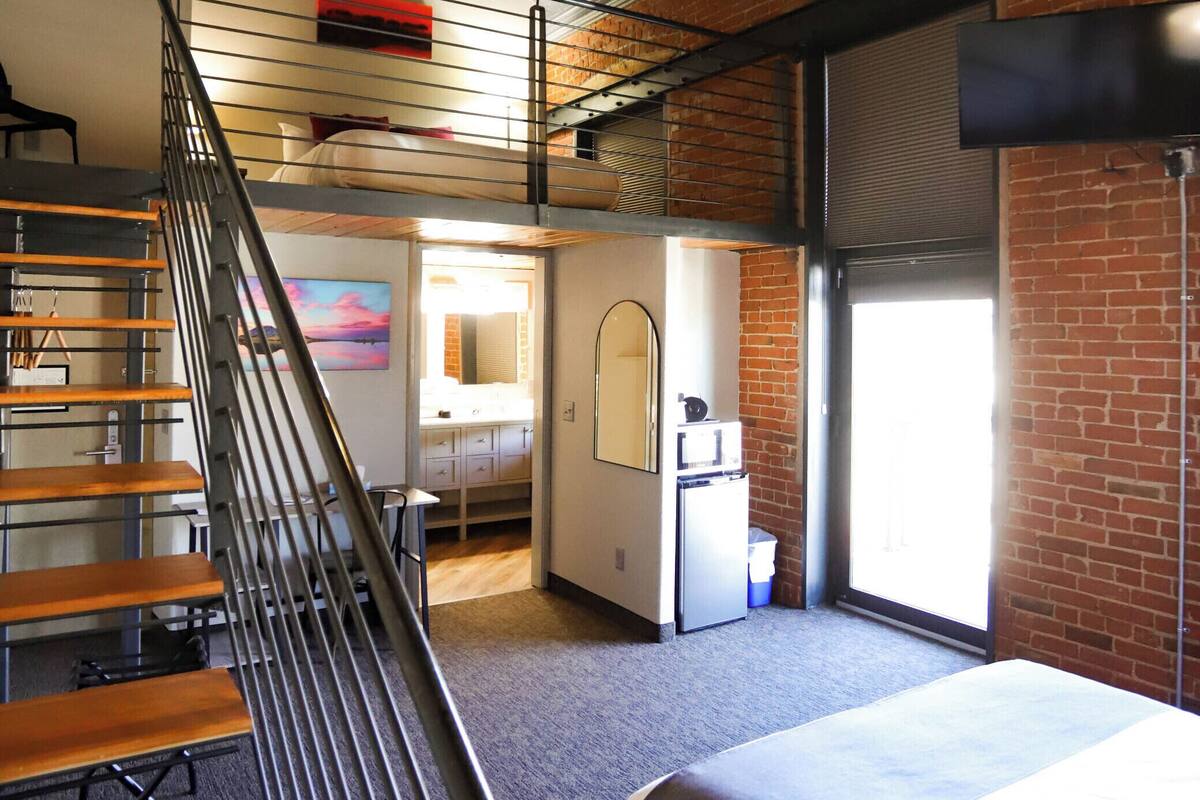 A hotel room with a bed, a desk, a chair, a small kitchenette, a bathroom, and a staircase.