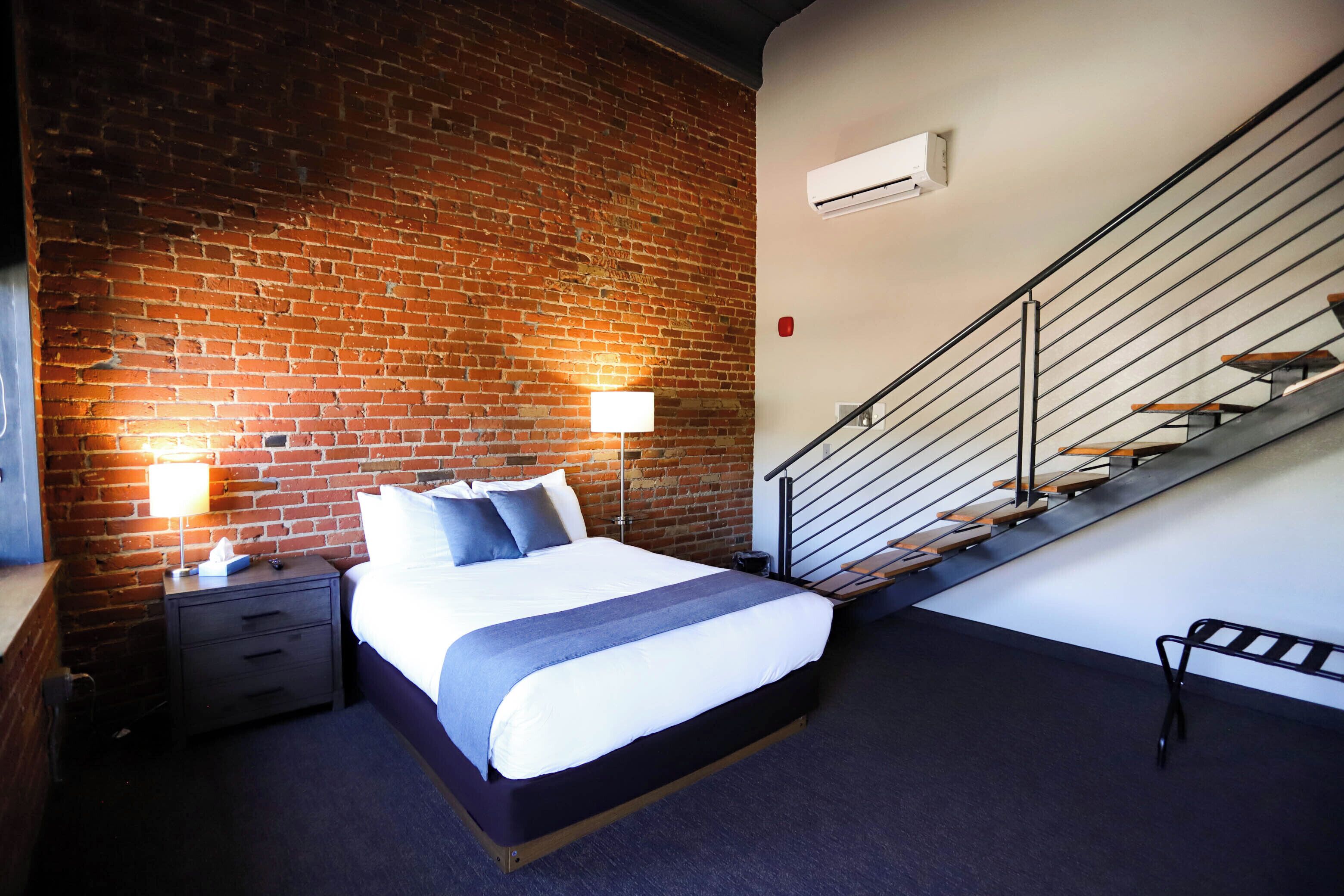 A hotel room with a brick wall, a bed with white bedding, a nightstand, a lamp, and a staircase with a metal railing.