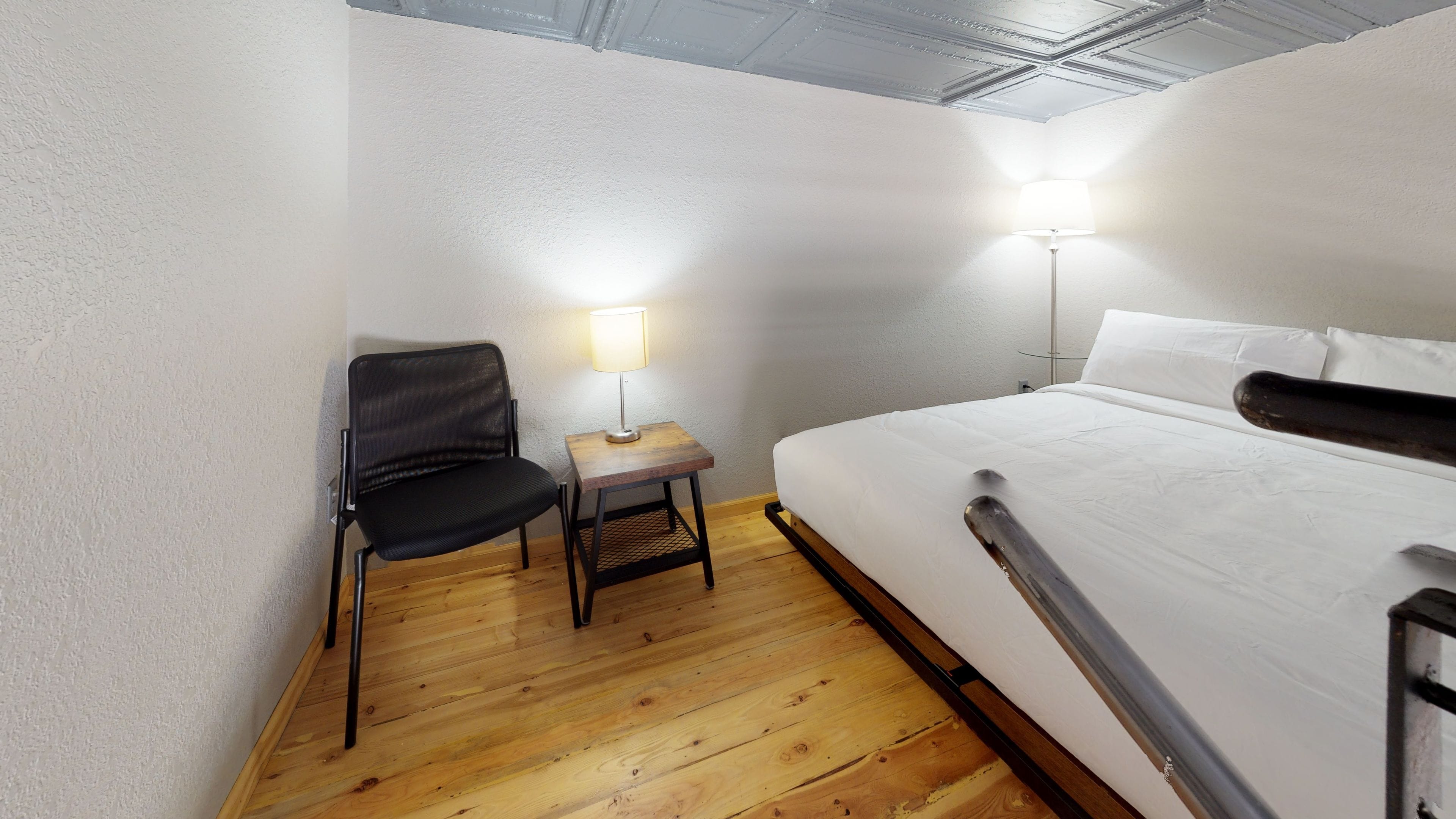 A hotel room with a bed, a chair, a small table with a lamp, and wooden flooring.