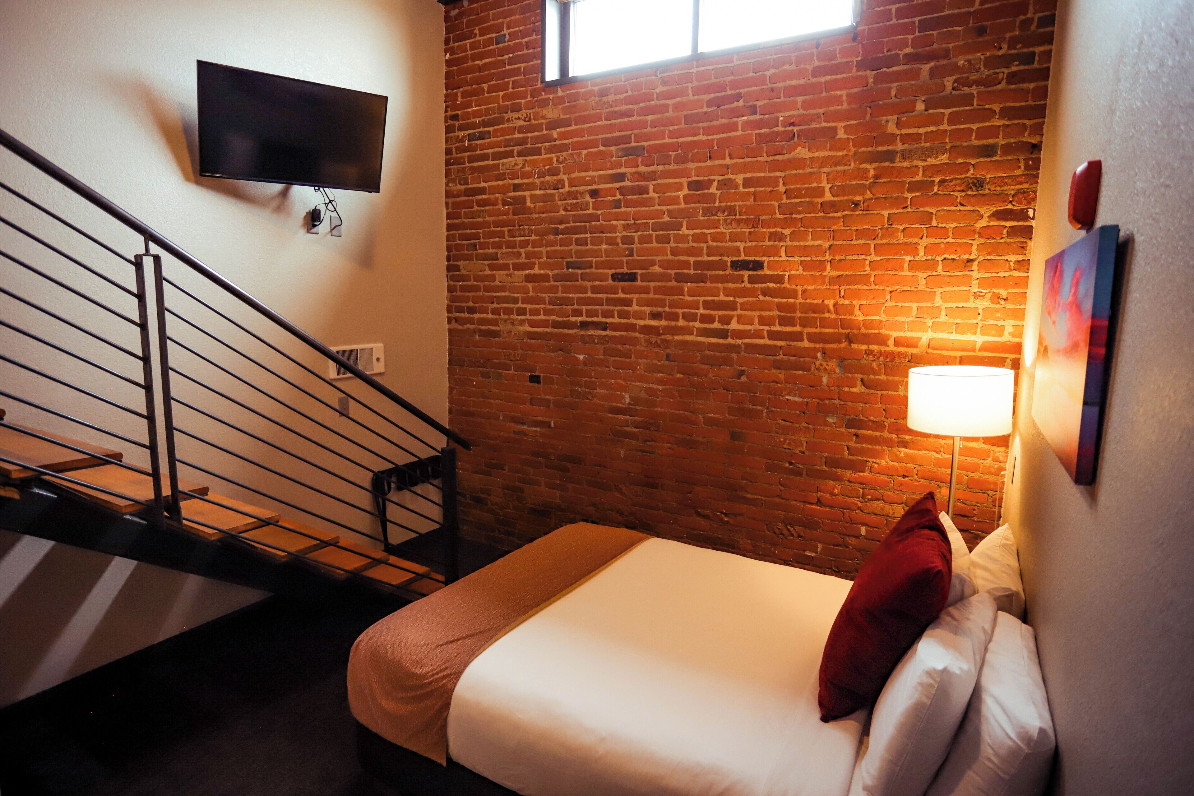 A hotel room with a brick wall, a flat-screen TV mounted above a staircase, a bed with white bedding and a red pillow, and a bedside lamp.
