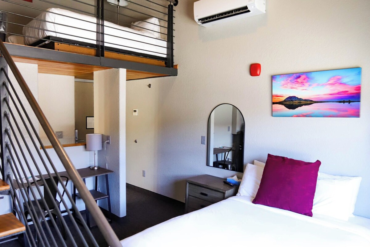 A hotel room with a bed, a desk, a chair, a mirror, an air conditioning unit, and a staircase.