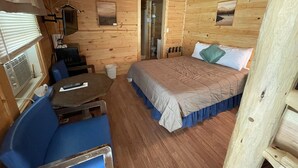 A wooden cabin room with a bed, a table, a chair, and a small desk.