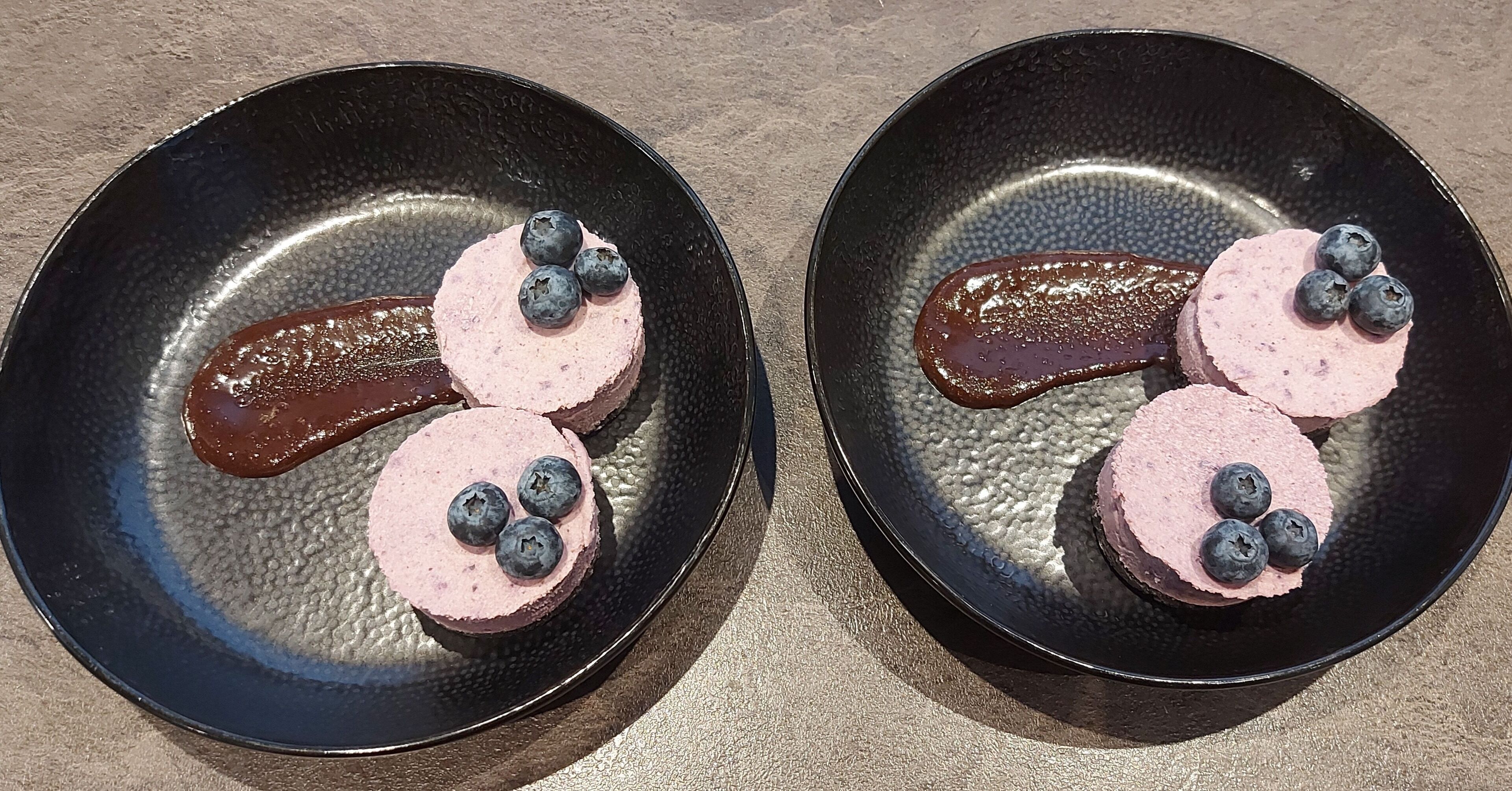 Two black plates with pink and chocolate desserts and blueberries on a textured surface.