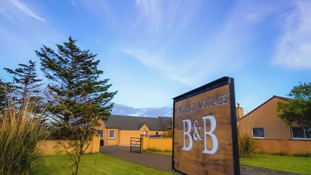 Señal que indica 'B&B', alojamiento tipo bed and breakfast, con un cielo despejado y una zona verde al frente.