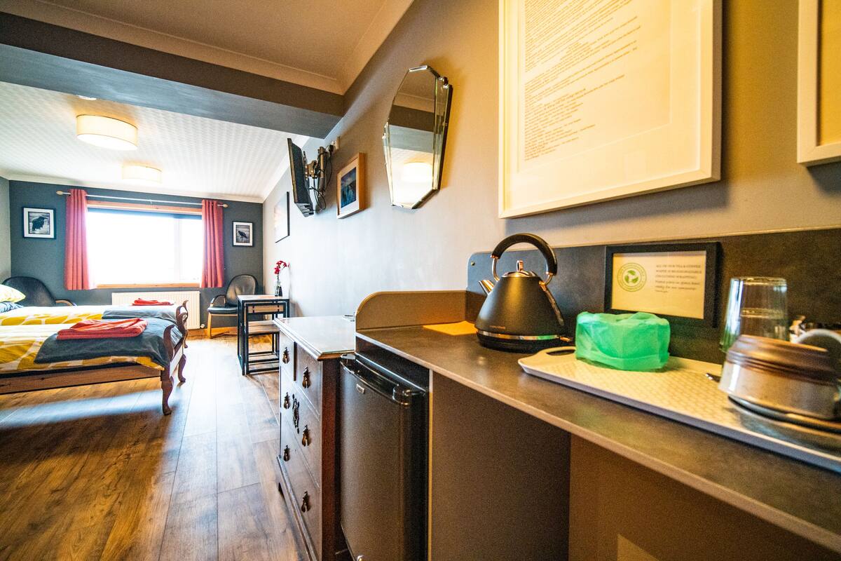 A hotel room with a wooden bed, a desk with a kettle, a green bag, and a framed certificate on the wall.