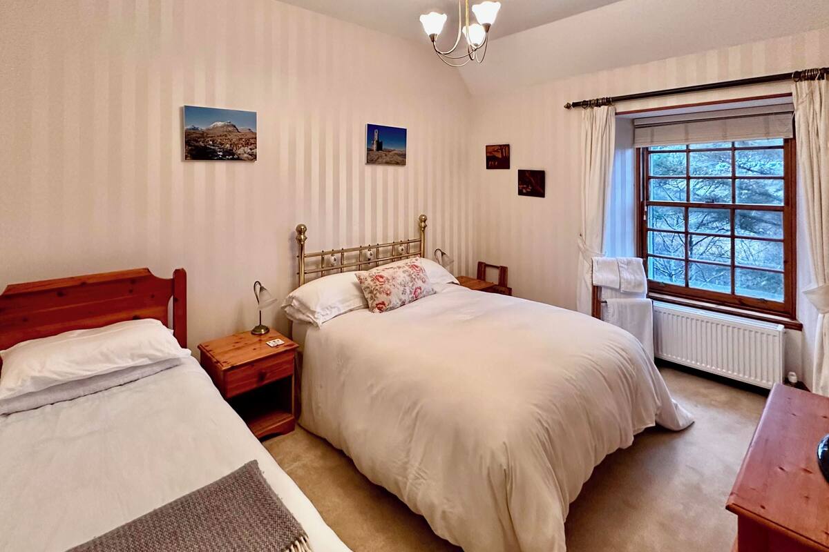 A bedroom with two single beds, a wooden headboard, a nightstand, a bedside lamp, a window with curtains, and a radiator.