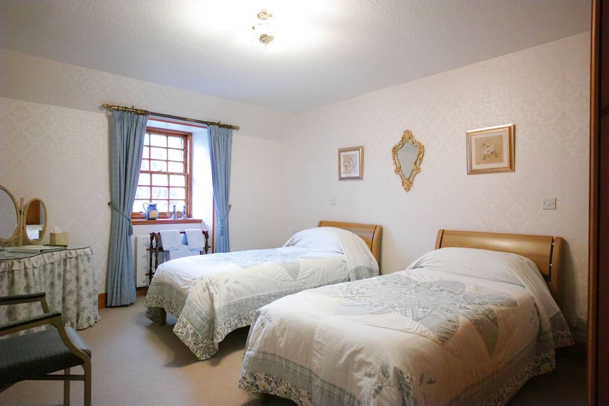 A bedroom with two single beds, a wooden chair, a desk with a mirror, and a window with curtains.