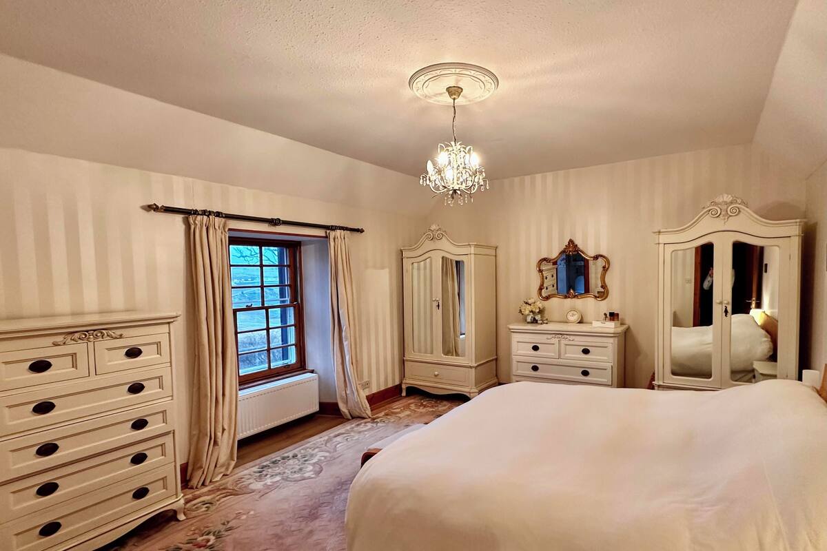 A bedroom with a large bed, a dresser, a mirror, and a window with curtains.