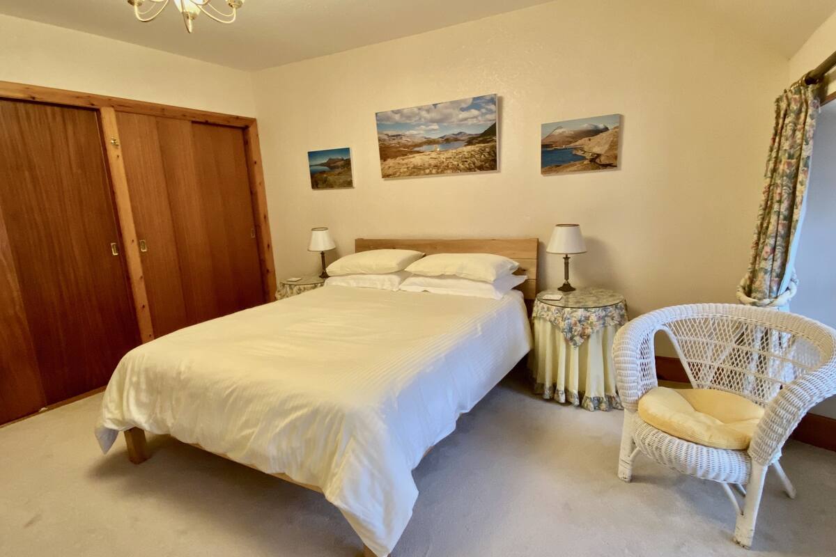 A bedroom with a large bed, two bedside tables with lamps, a wicker chair, and three landscape paintings on the wall.