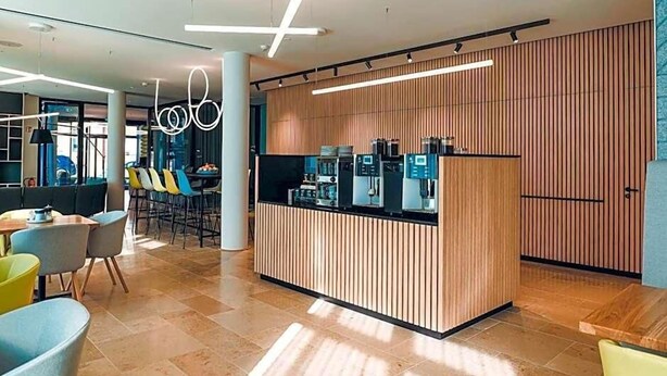 A modern reception area with a wooden panel wall, a coffee station, and seating arrangements. - Art Hotel Ahlen (Ahlen)
