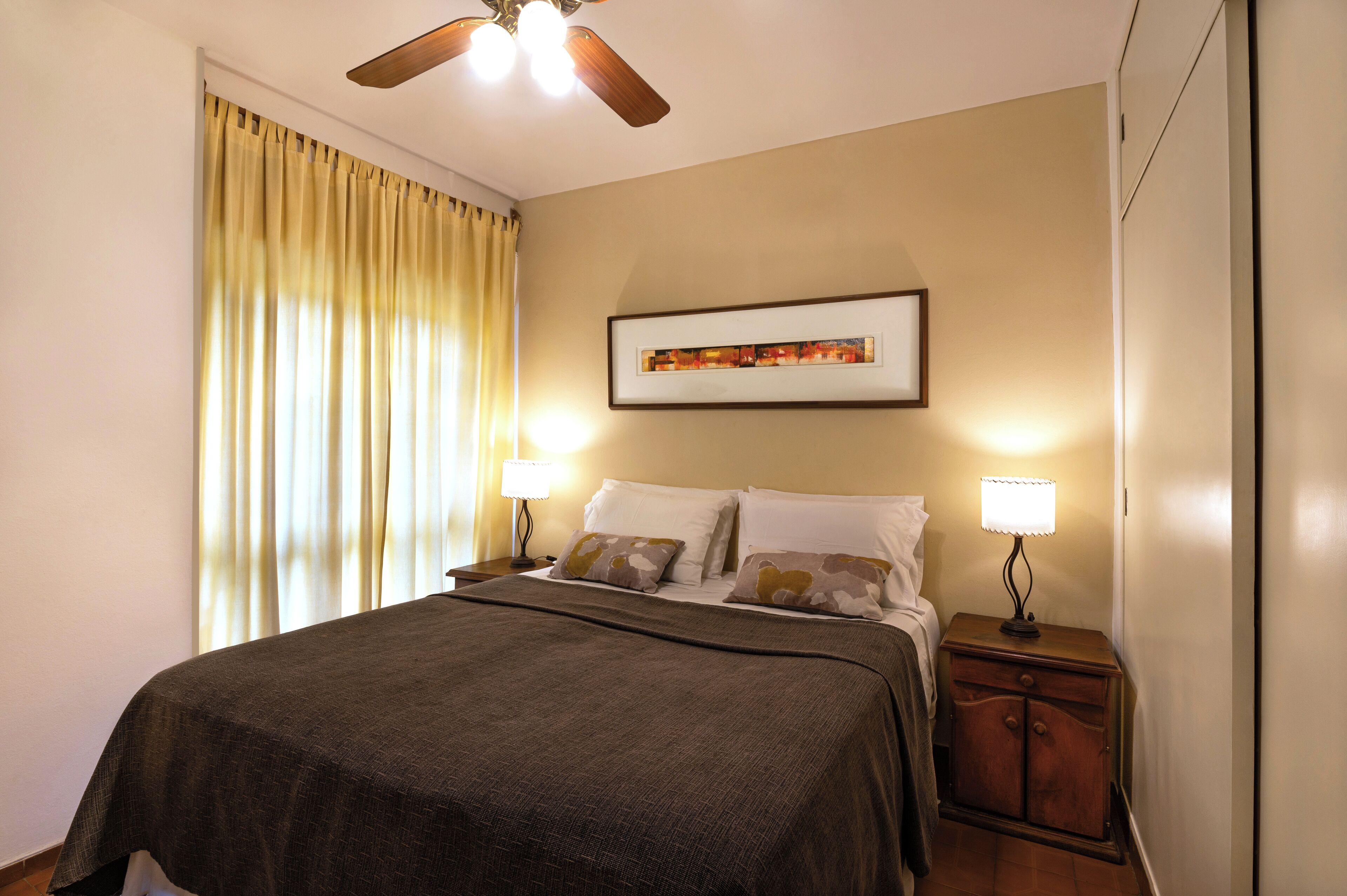 A neatly made bed with a dark bedspread, two bedside lamps, a ceiling fan, and a framed picture on the wall.